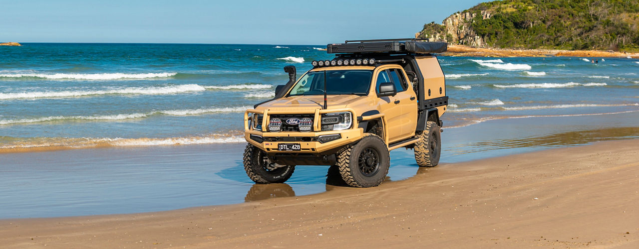 Ford Ranger Super Duty fitted with a MITS Alloy canopy, rooftop tent and awning, parked on a sandy beach beside the ocean.