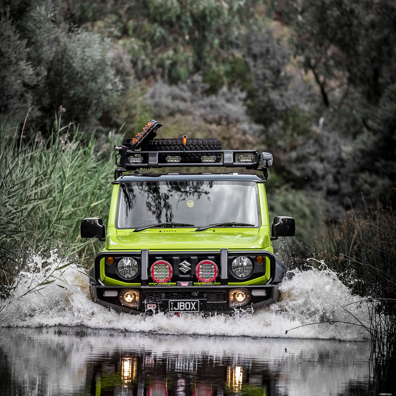 Front view of an ARB equipped Suzuki Jimny crossing a creek with driving lights illuminated and water splashing around the vehicle.