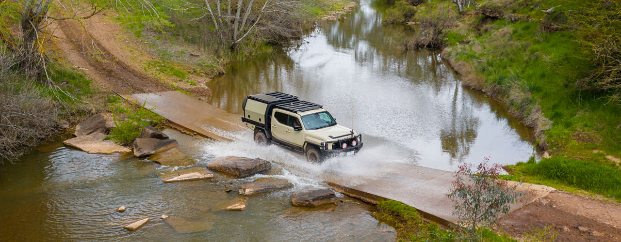 Aerial view of a Kia Tasman fitted with a MITS Alloy canopy crossing a river causeway, with water splashing around the vehicle in a rural landscape.