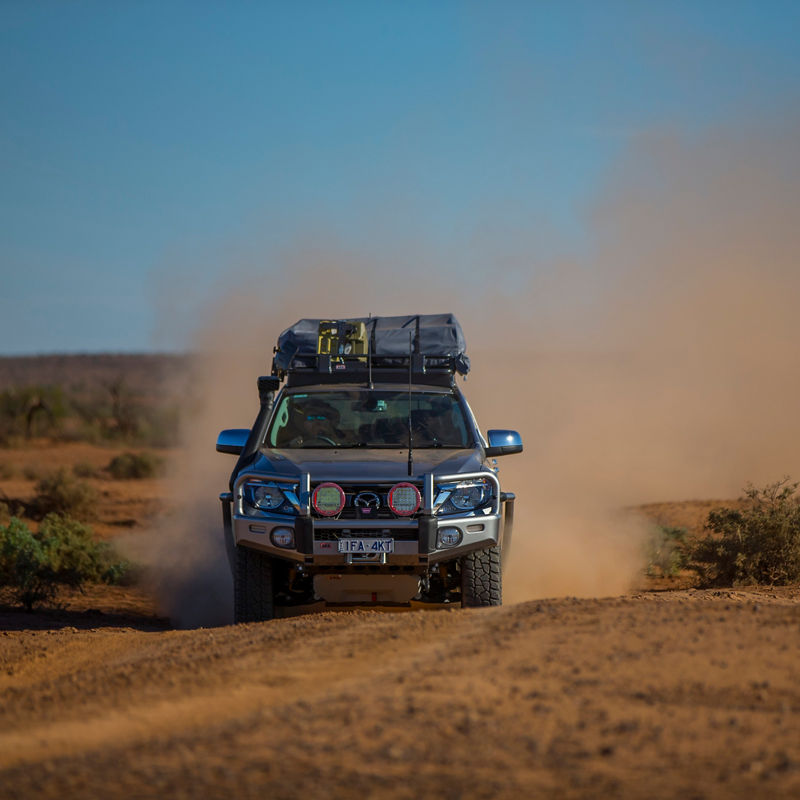 Front view of an ARB equipped Mazda BT-50 driving through a dusty desert landscape with a roof rack system loaded with touring gear.