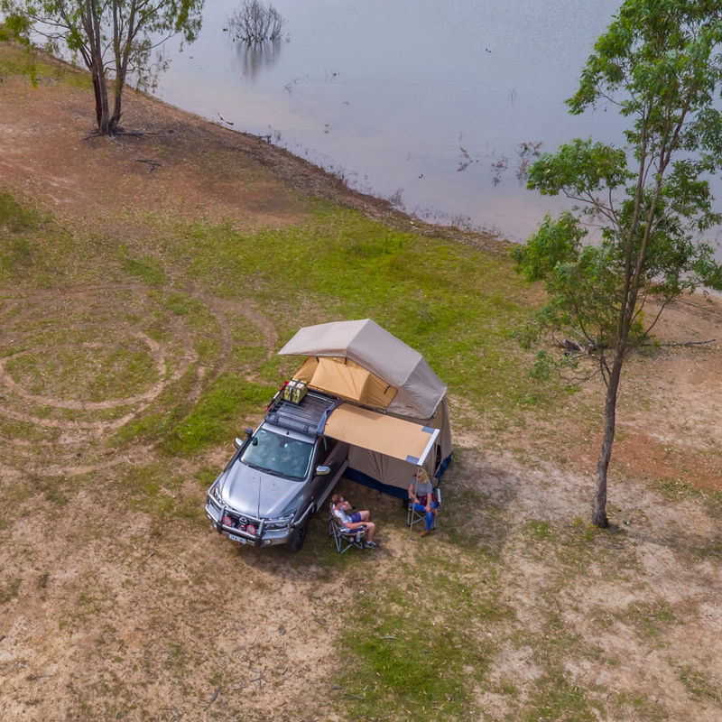 Aerial view of an ARB equipped Mazda BT-50 set up at a lakeside campsite with a rooftop tent and awning, while two people relax beside the vehicle.
