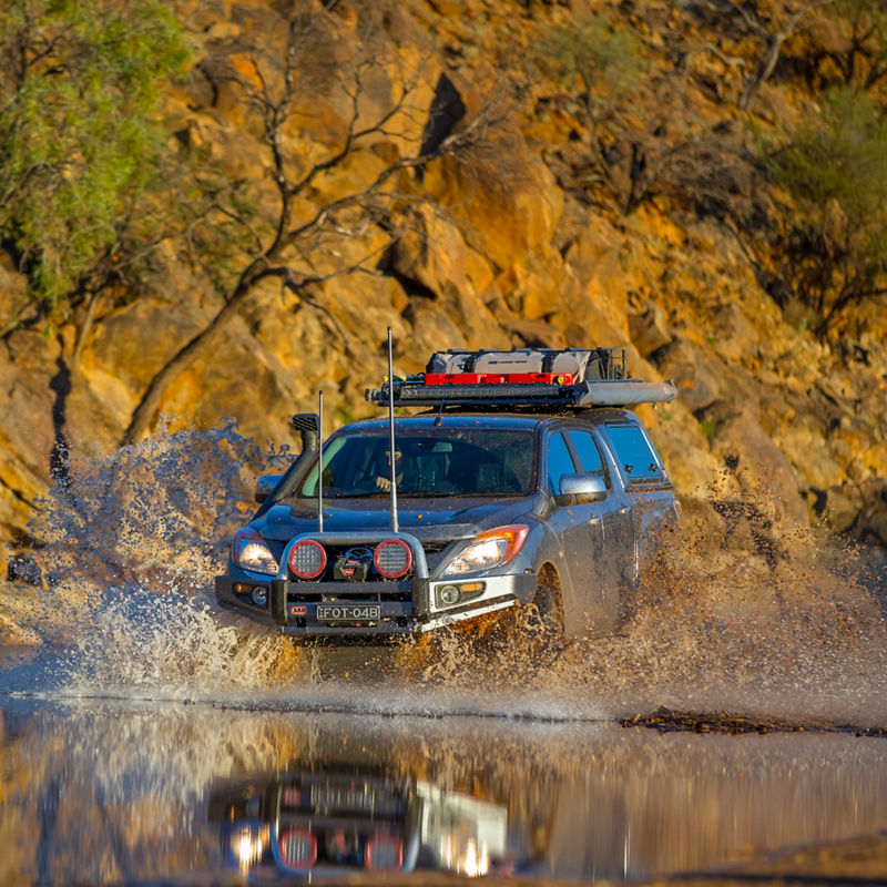 ARB equipped Mazda BT-50 crossing a creek with water splashing around the vehicle in a rocky outback landscape.