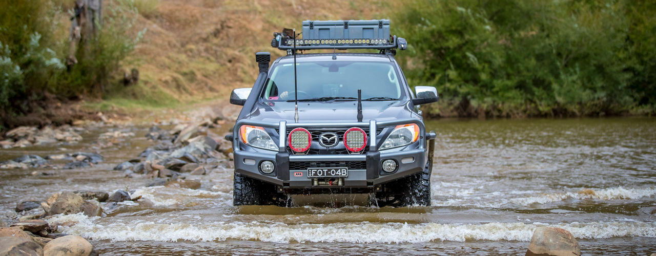 Front view of a Mazda BT-50 crossing a shallow rocky river, fitted with an ARB bull bar, driving lights, snorkel, roof rack system and recovery gear.
