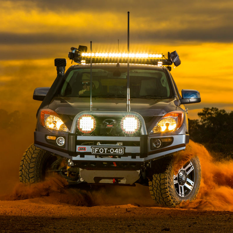 Front view of an ARB equipped Mazda BT-50 driving through sand at sunset with driving lights and a roof mounted light bar illuminated.