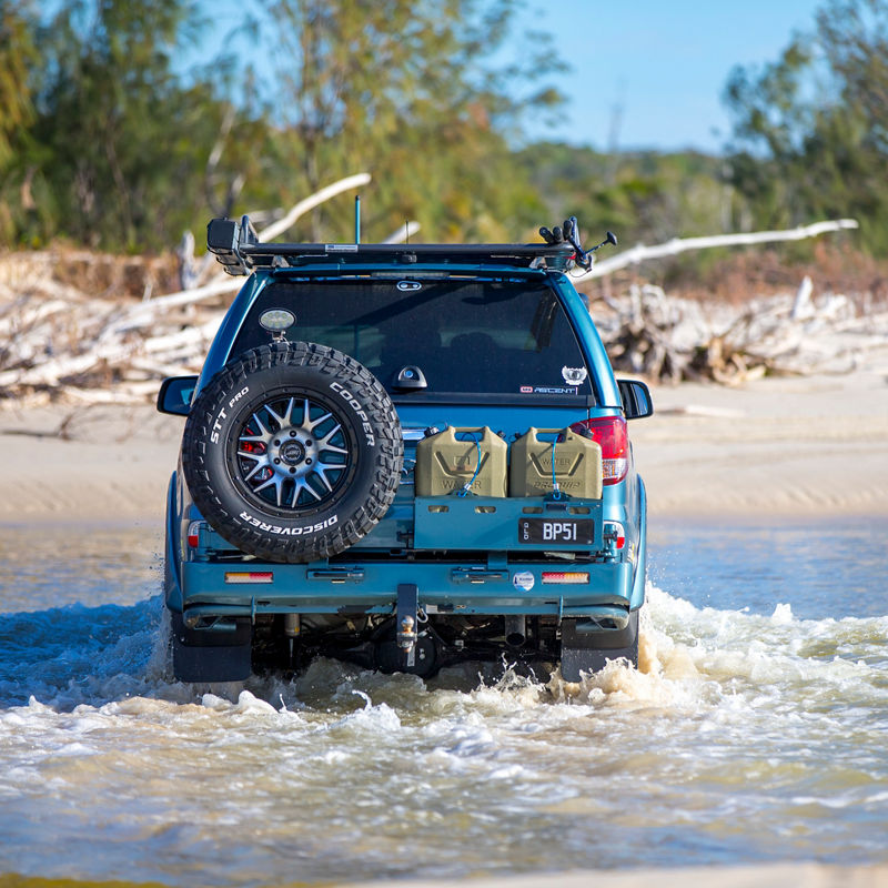 Rear view of an ARB equipped Mazda BT-50 crossing a shallow coastal creek with a rear wheel carrier, jerry cans and roof rack system.