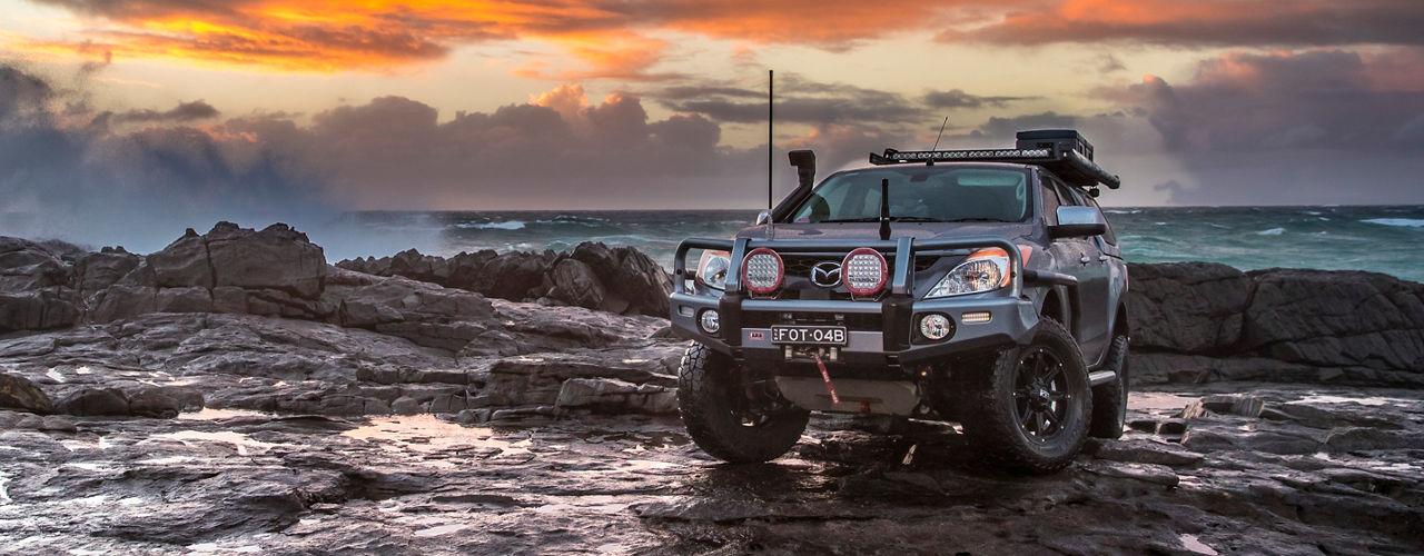 Mazda BT-50 fitted with an ARB bull bar, driving lights, snorkel, roof rack system and recovery gear, parked on a rocky coastal shoreline beneath a dramatic sunset sky.