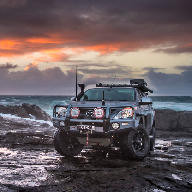ARB equipped Mazda BT-50 parked on a rocky coastal shoreline at sunset with a bull bar, driving lights, roof rack system and canopy.