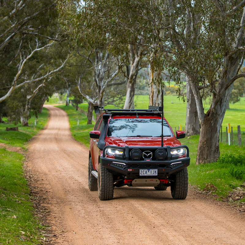 ARB equipped Mazda BT-50 driving along a winding gravel road through a rural landscape lined with gum trees.