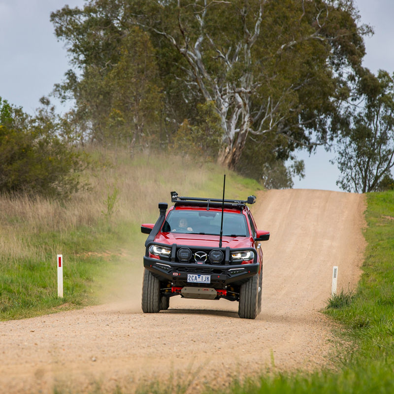 Front view of an ARB equipped Mazda BT-50 driving along a gravel road through a rural landscape with dust trailing behind the vehicle.