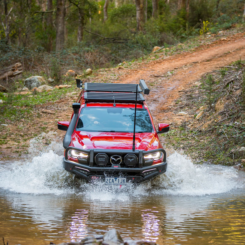Front view of an ARB equipped Mazda BT-50 crossing a creek with water splashing around the vehicle in a forested off road environment.