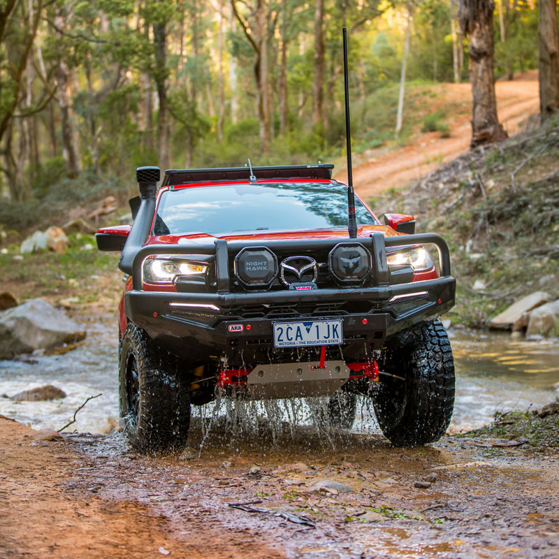 Front view of an ARB equipped Mazda BT-50 crossing a shallow creek with water dripping from the vehicle, fitted with a bull bar, driving lights and under vehicle protection.