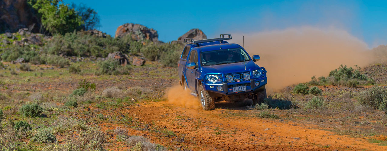 Front view of a Mitsubishi Triton driving along a dusty outback track, fitted with an ARB bull bar, canopy and roof rack system, with dust trailing behind the vehicle.