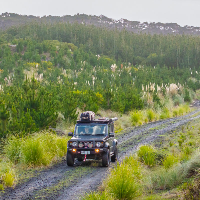 ARB equipped Suzuki Jimny driving along a gravel track through a forested landscape.