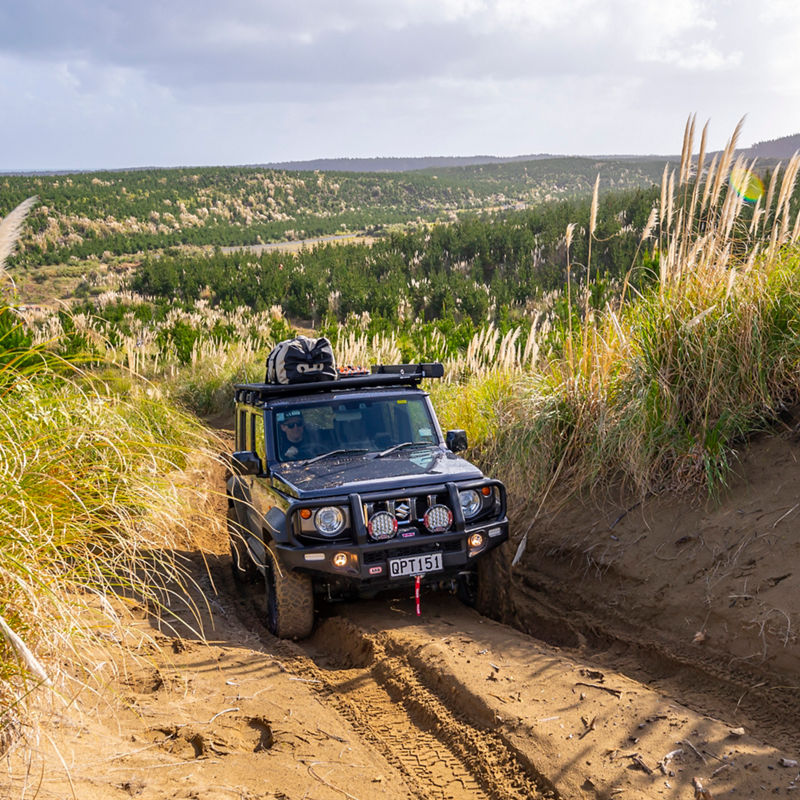 ARB equipped Suzuki Jimny driving through deep sand ruts in a coastal dune environment.