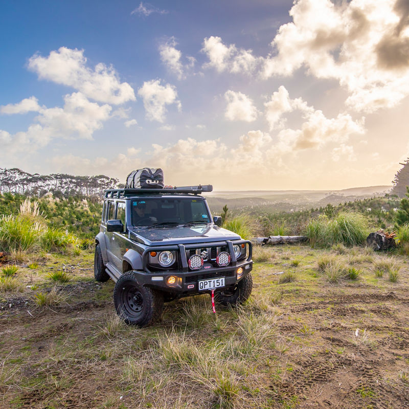 ARB equipped Suzuki Jimny parked in a remote landscape with a roof rack system, under a dramatic sky at sunset.