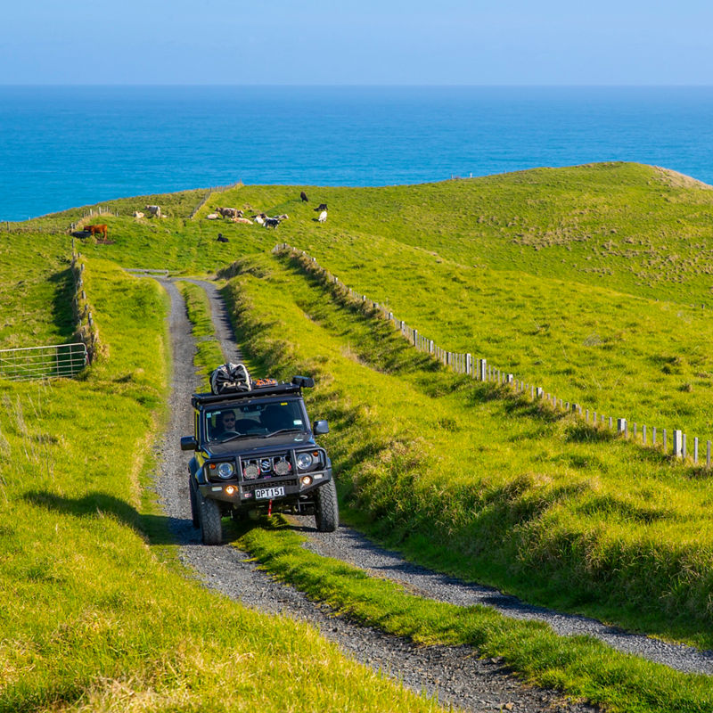 ARB equipped Suzuki Jimny driving along a gravel track through rolling coastal farmland with the ocean in the background.