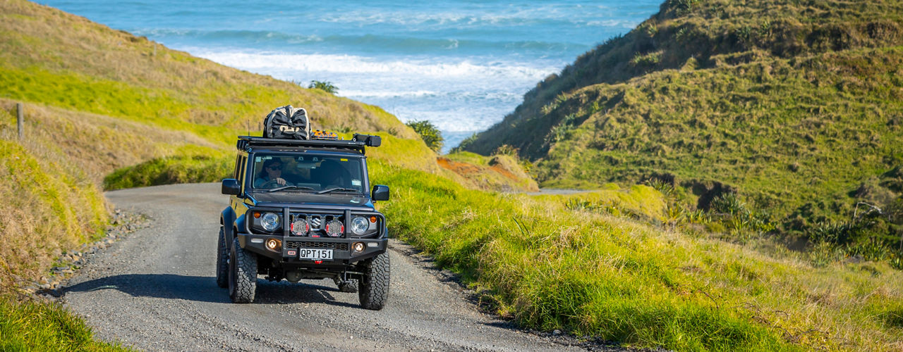 Suzuki Jimny driving along a winding gravel road through a coastal landscape, fitted with an ARB bull bar, driving lights, roof rack system and touring gear.