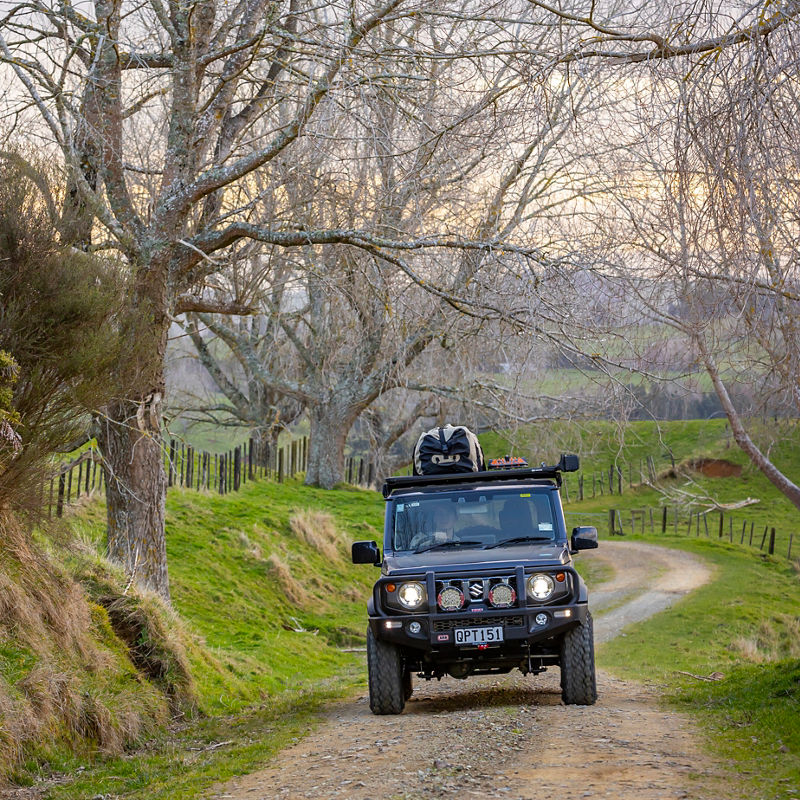 ARB equipped Suzuki Jimny driving along a winding gravel road through a rural landscape with bare trees and rolling farmland.