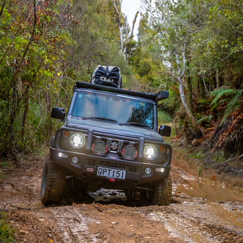 Front view of an ARB equipped Suzuki Jimny driving along a muddy forest track with driving lights illuminated.