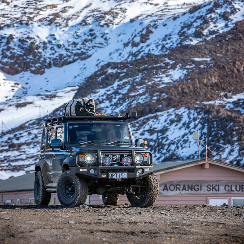 ARB equipped Suzuki Jimny parked in front of snow covered mountains near Aorangi Ski Club, fitted with a roof rack system and recovery equipment.