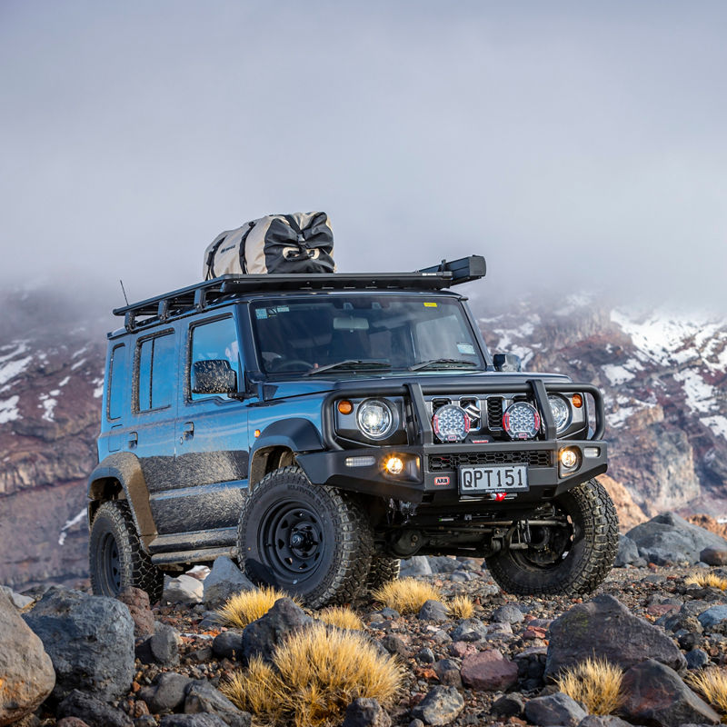 ARB equipped Suzuki Jimny fitted with a Summit Bull Bar, driving lights and roof rack system parked on rocky terrain with snow covered mountains in the background.