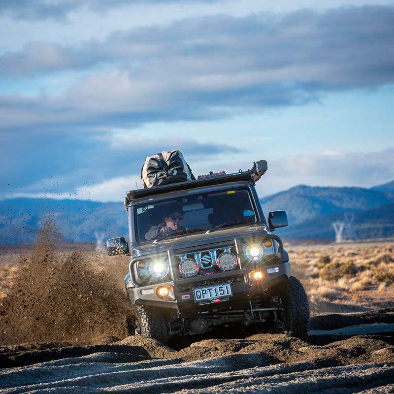 ARB equipped Suzuki Jimny driving through deep sand with driving lights illuminated and sand spraying from the front wheels, against a mountain backdrop.