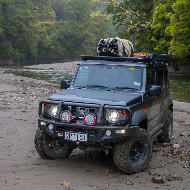 ARB equipped Suzuki Jimny fitted with a roof rack system and rooftop tent on a rocky riverbed surrounded by dense rainforest.