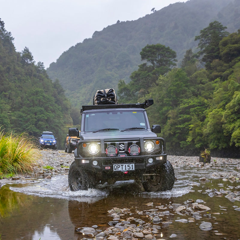 Front view of an ARB equipped Suzuki Jimny crossing a rocky creek with driving lights illuminated, surrounded by dense rainforest and mountains.