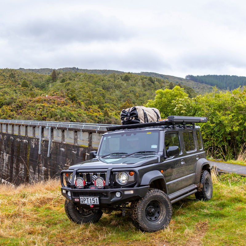 ARB equipped Suzuki Jimny parked beside a dam in a forested mountain landscape with a roof rack system and awning.