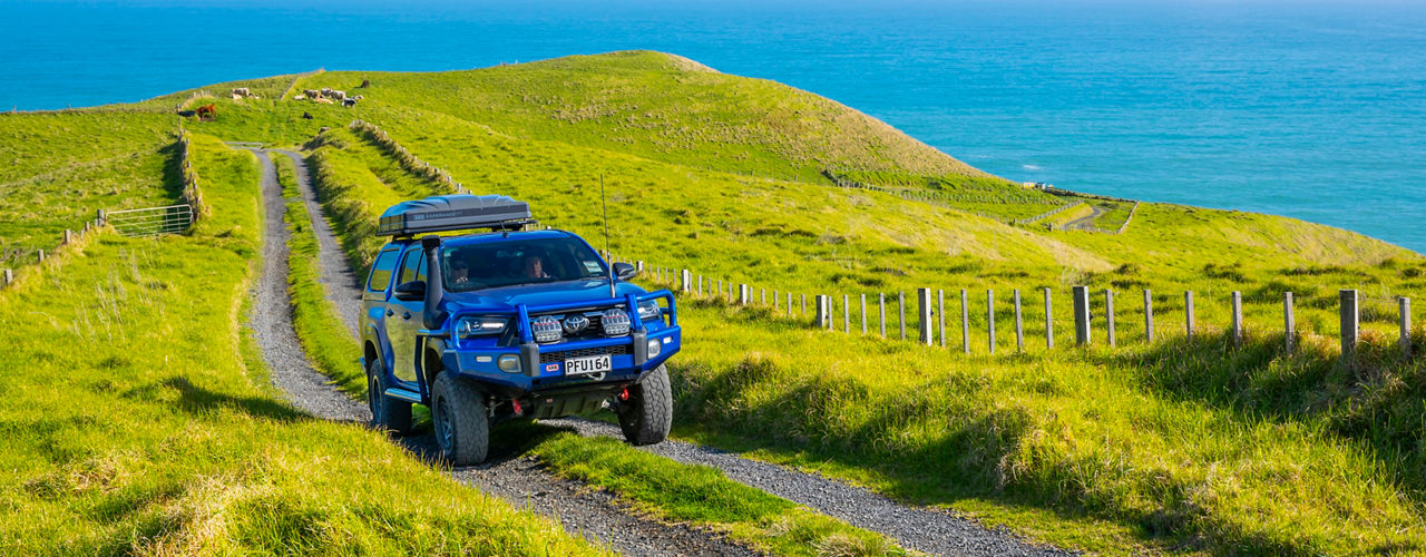 ARB equipped Toyota HiLux with a canopy and roof rack driving along a gravel track through coastal farmland.