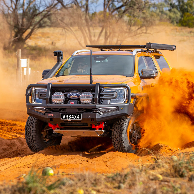 Front view of a Ford Ranger fitted with an ARB bull bar, driving lights, snorkel, roof rack and awning, driving through deep red sand.