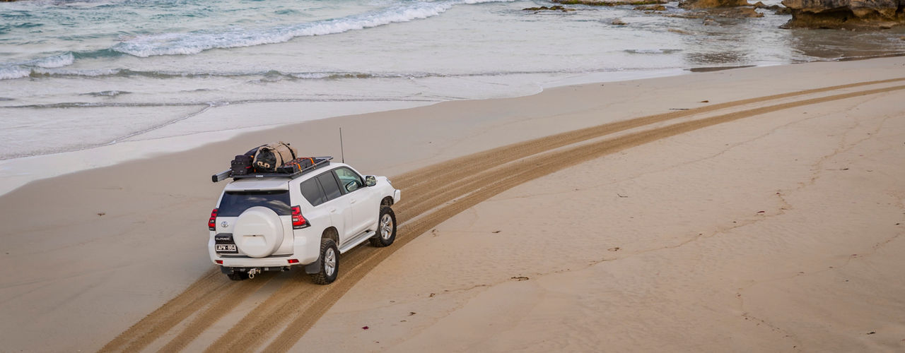 Aerial rear three quarter view of a Toyota LandCruiser Prado 150 Series driving along a sandy beach beside the ocean, fitted with an ARB BASE Rack carrying touring gear.