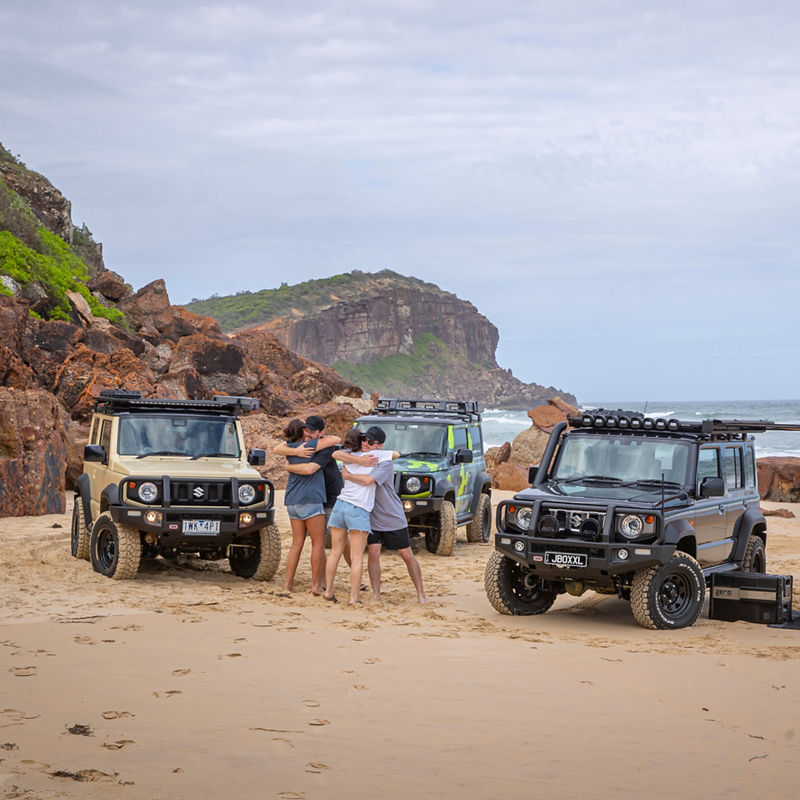 Group of people standing together on a beach beside three ARB equipped Suzuki Jimnys, with rocky coastal cliffs in the background.