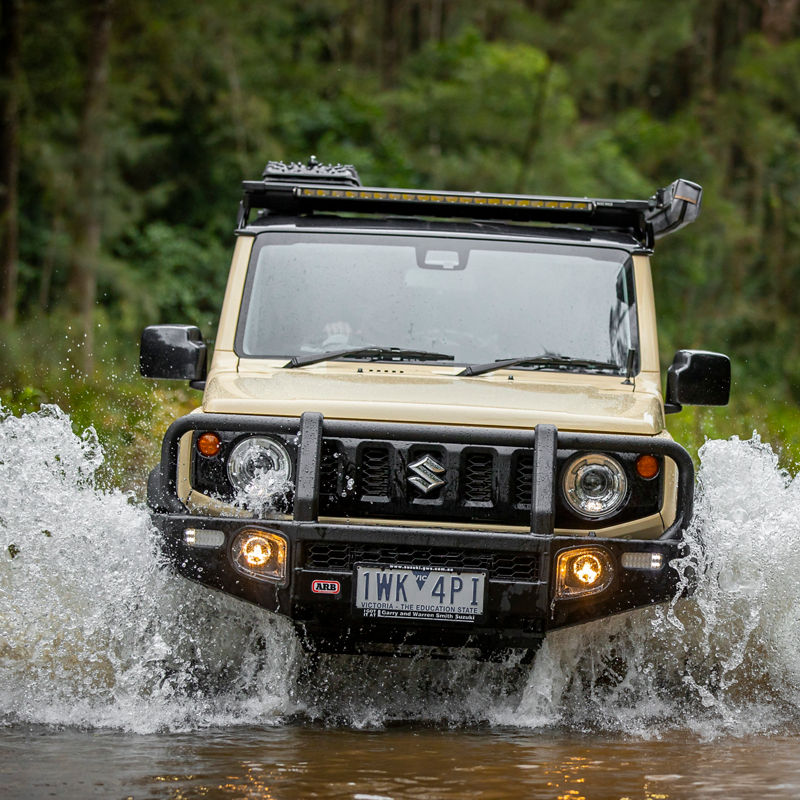 Front view of an ARB equipped Suzuki Jimny crossing a creek with water splashing around the vehicle in a forested off road environment.