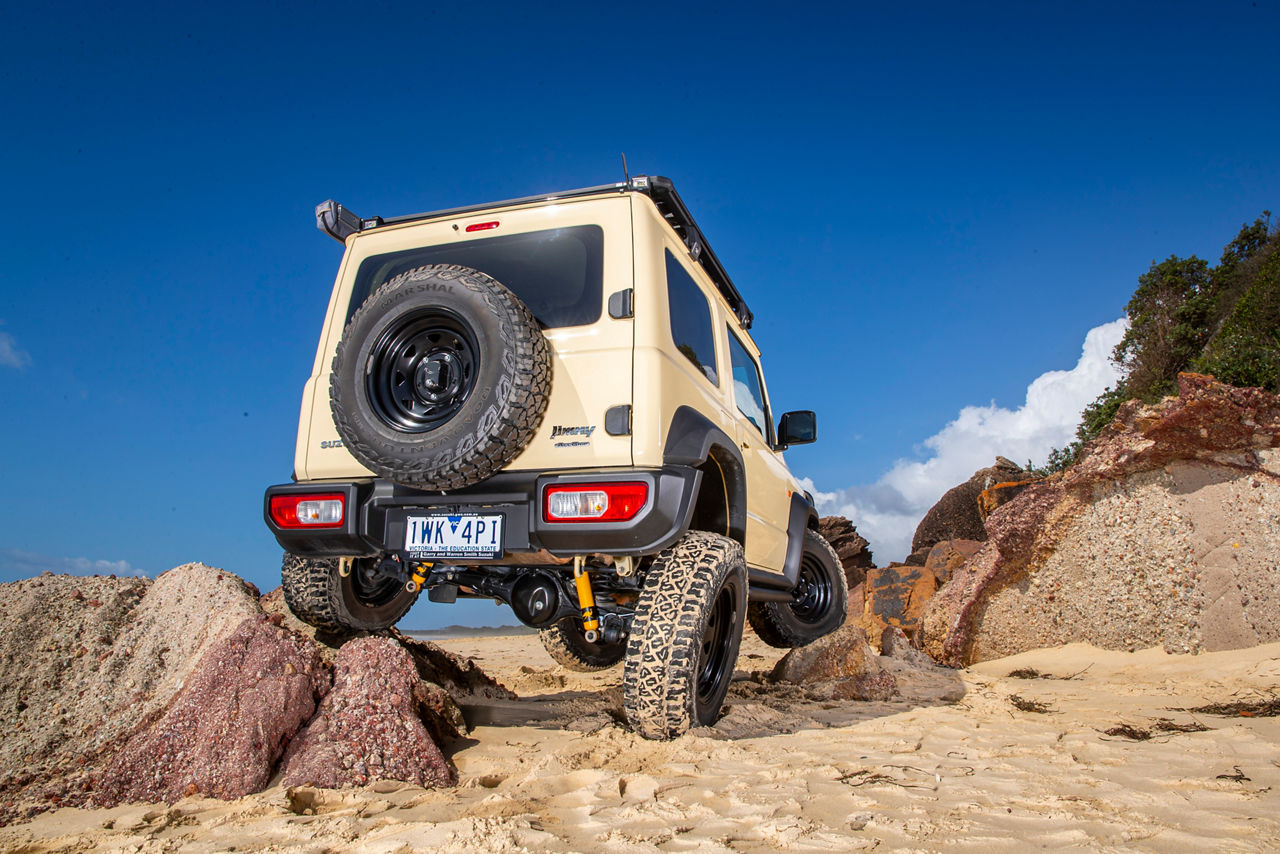 Rear view of a Suzuki Jimny navigating rocky terrain with Old Man Emu Nitrocharger suspension components visible beneath the vehicle.