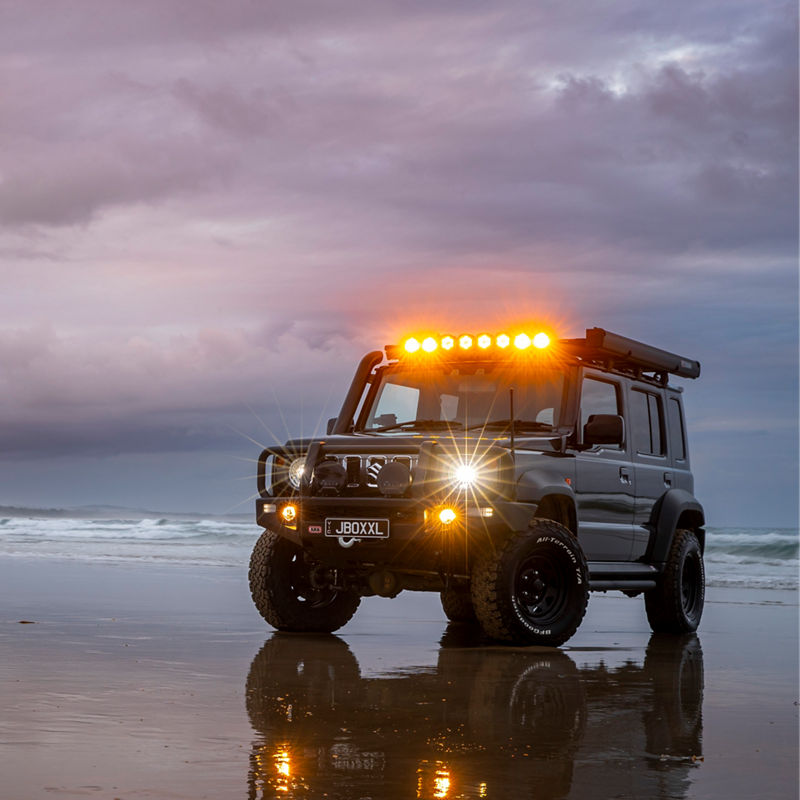 ARB equipped Suzuki Jimny parked on a beach at dusk with NACHO Quatro driving lights illuminated and reflected on wet sand.