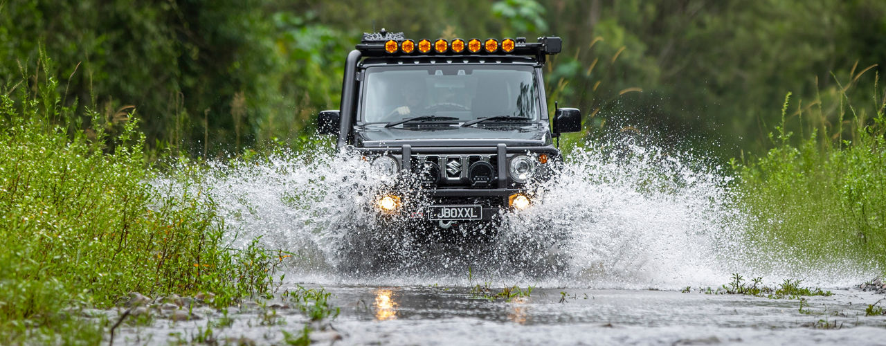 Front view of a Suzuki Jimny crossing a flooded track, fitted with an ARB bull bar, roof mounted driving lights and snorkel, with water splashing around the vehicle.