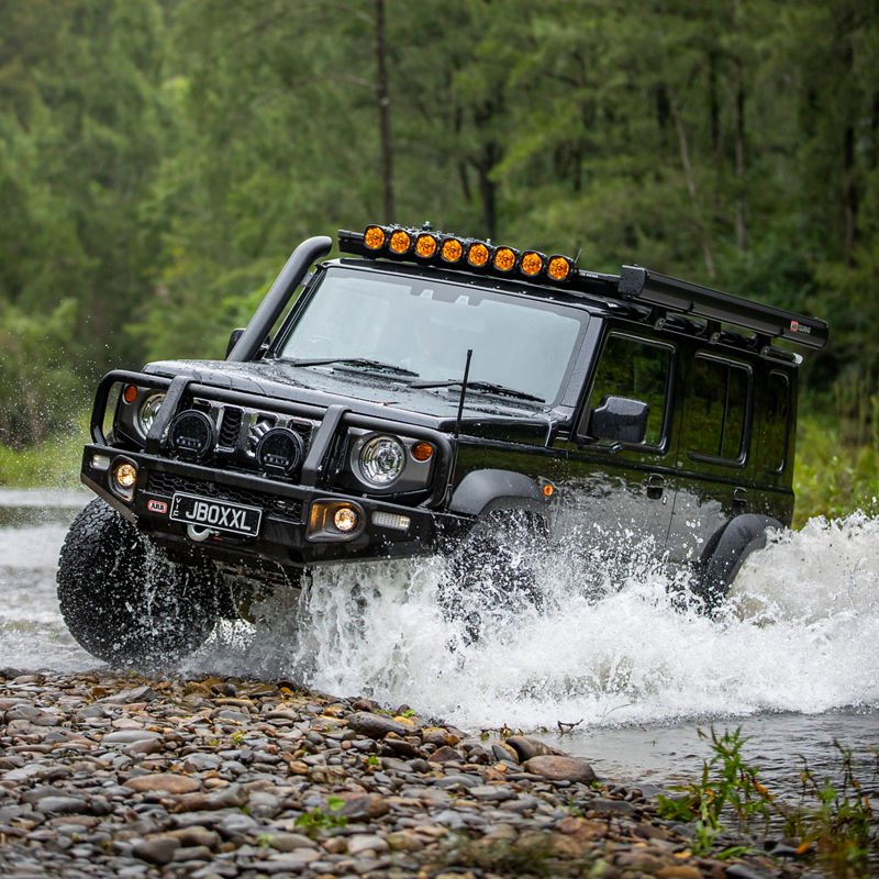 ARB equipped Suzuki Jimny crossing a rocky creek with water splashing around the vehicle, fitted with driving lights, a roof rack system and awning.