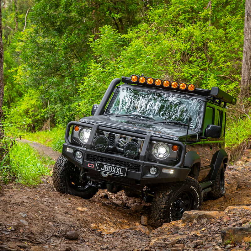 ARB equipped Suzuki Jimny navigating a rocky forest track with NACHO Quatro driving lights illuminated and visible wheel articulation.