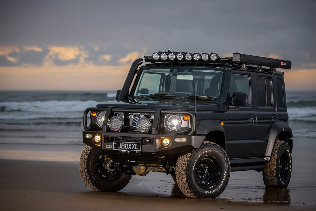 ARB equipped Suzuki Jimny fitted with a Summit Bull Bar, driving lights and roof rack system parked on a beach at dusk.