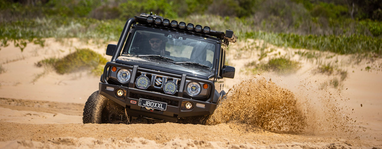 ARB equipped Suzuki Jimny driving through deep sand with driving lights illuminated and sand spraying from the front wheels.