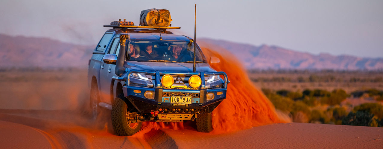 Mitsubishi Triton fitted with an ARB bull bar, driving lights, canopy and roof rack system driving through sand dunes at sunset.