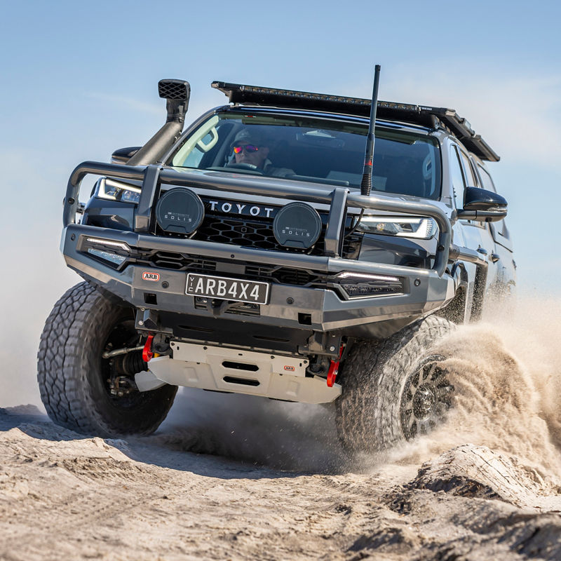 Front three-quarter view of a Toyota HiLux fitted with an ARB Bull Bar, driving lights, snorkel, roof rack and under-vehicle protection, driving through deep sand.