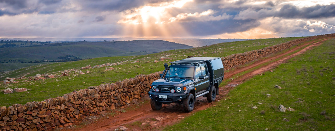 Toyota LandCruiser 79 Series fitted with a tray canopy and roof rack system, driving along a rural dirt road beside a stone wall beneath dramatic sunset clouds.