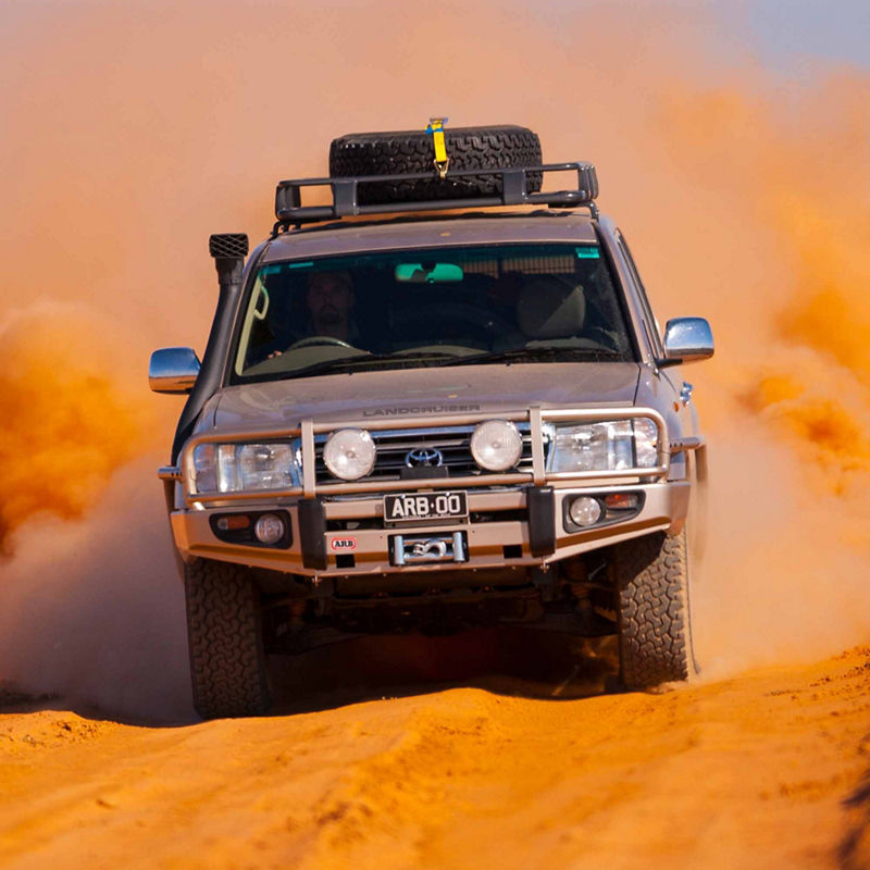 Front view of a Toyota LandCruiser 100 Series driving through deep sand, fitted with an ARB bull bar, driving lights, roof rack system, snorkel and winch, with dust trailing behind the vehicle.