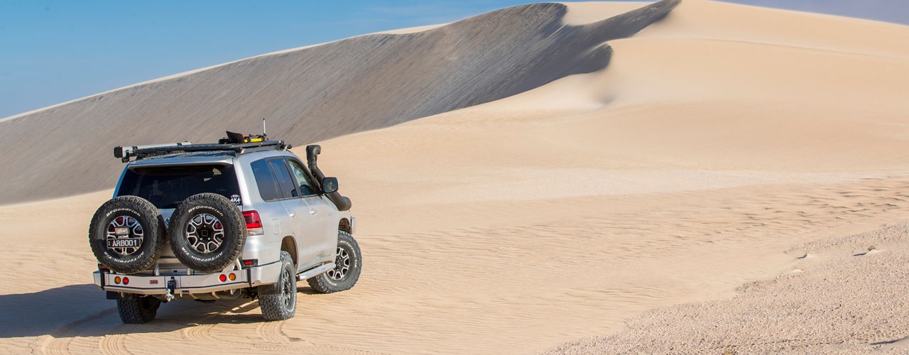 Rear three quarter view of a Toyota LandCruiser 200 Series driving through sand dunes, fitted with a roof rack system, awning, snorkel and rear bar with dual spare wheel carriers.