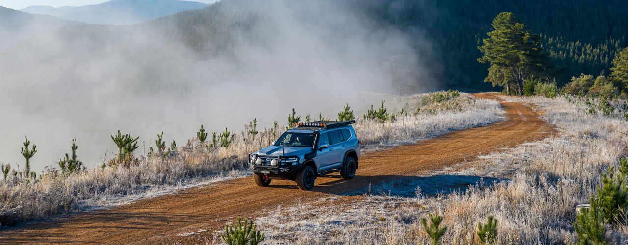ARB equipped Toyota LandCruiser 300 Series driving along a winding gravel road through a frosted mountain landscape with mist rising from the valley below.