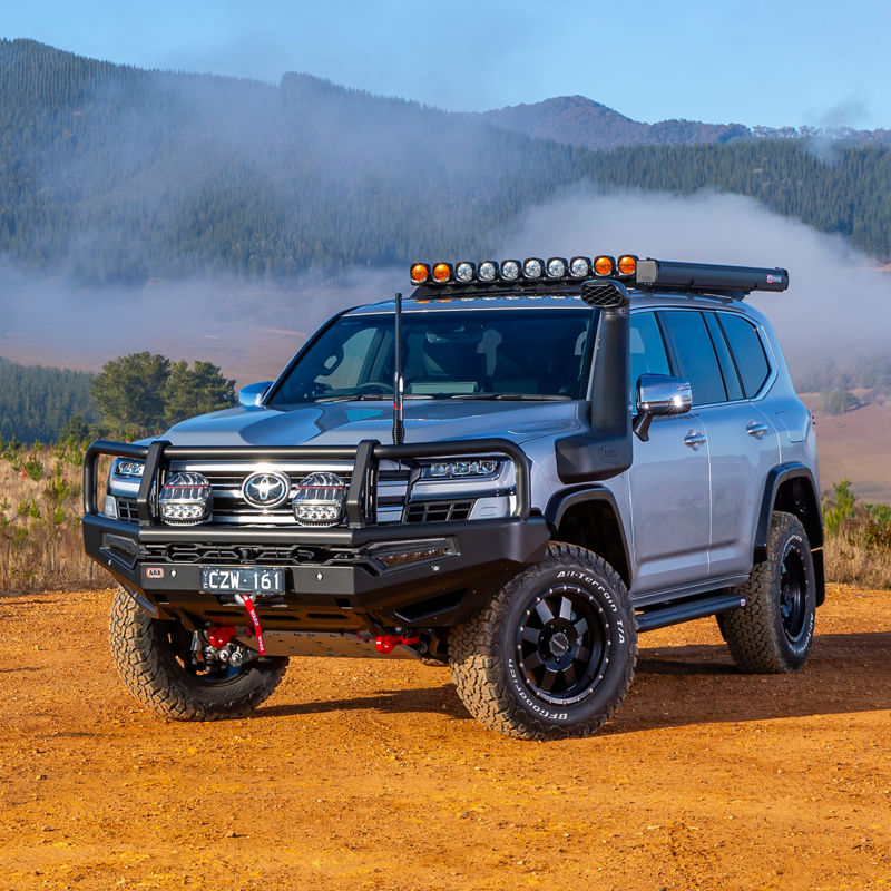 Toyota LandCruiser 300 Series fitted with an ARB bull bar, driving lights, roof rack system and snorkel, parked on a gravel track overlooking a misty mountain landscape.