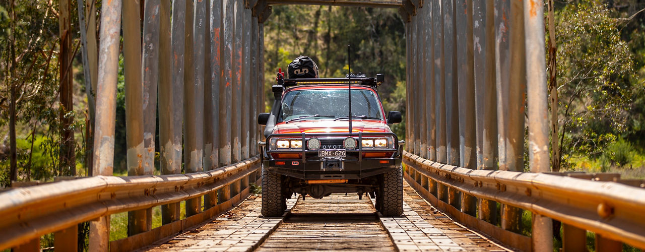 Front view of a Toyota LandCruiser 80 Series crossing a historic timber and steel bridge, fitted with an ARB bull bar, driving lights, roof rack system and auxiliary lighting.