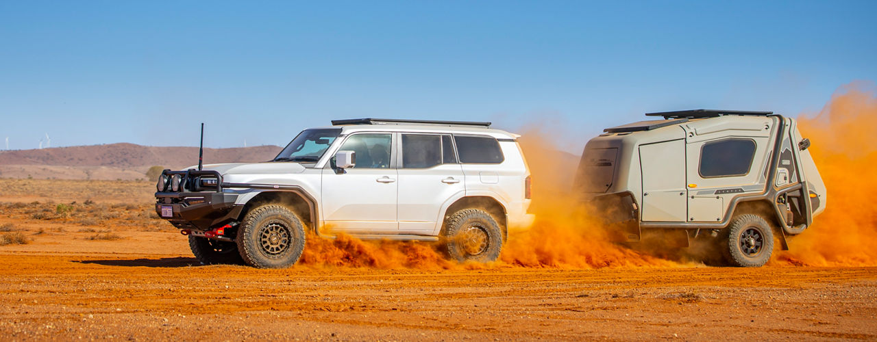 Side view of a Toyota Prado 250 fitted with an ARB bull bar, roof rack and off-road tyres, towing an ARB camper trailer across red desert terrain.